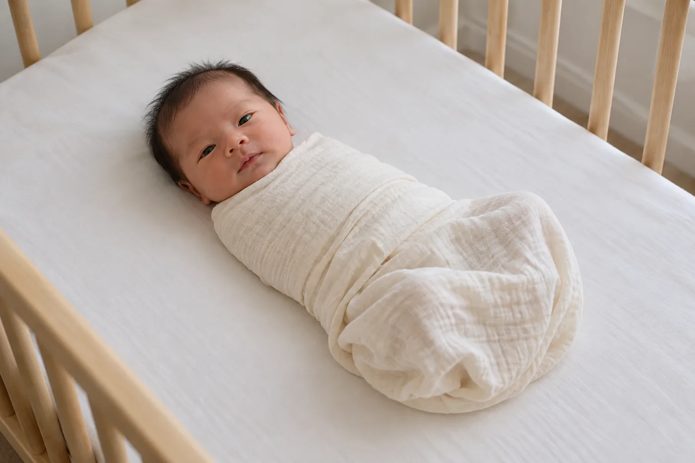Bé quấn khăn mỏng nằm ngửa trên ga phẳng trong nôi.